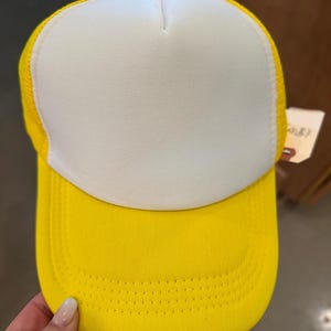 Puede incluir: Una gorra de camionero amarilla y blanca con una parte trasera de malla. La parte delantera de la gorra es blanca lisa y la visera es amarilla.