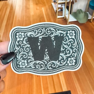 May include: A black and white decorative sticker in a western style. The sticker features a black letter "W" in the centre, surrounded by a floral and scroll design. The border has a rope-like pattern.