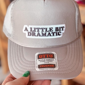 Op de afbeelding: Een grijze trucker-pet met een zwart-witte geborduurde patch met de tekst "A Little Bit Dramatic". De pet heeft een zwart-wit label met de tekst "Otto World Famous Foam Front OSFM Trucker Durable Snap Closure".