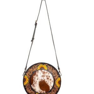 May include: A round, brown leather crossbody bag with a white and brown cowhide patch in the center. The bag is decorated with yellow sunflowers and intricate floral designs.