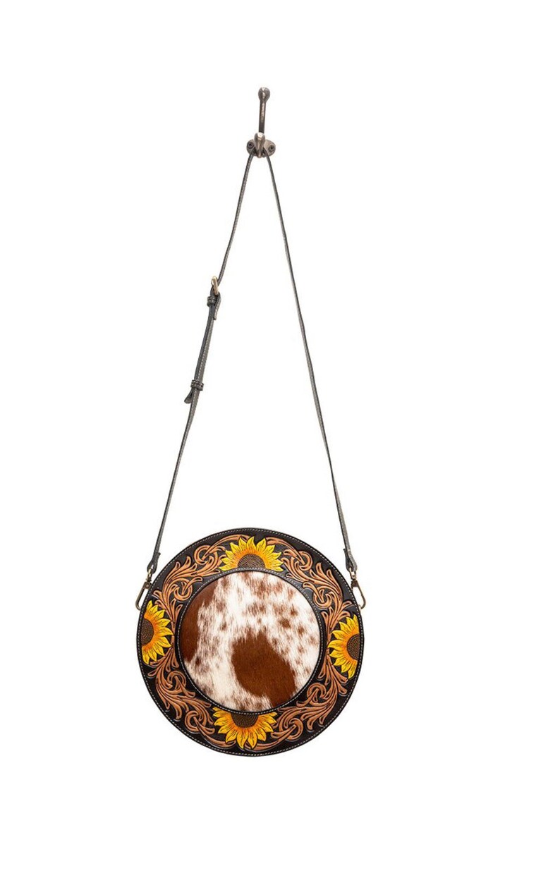 May include: A round, brown leather crossbody bag with a white and brown cowhide patch in the center. The bag is decorated with yellow sunflowers and intricate floral designs.
