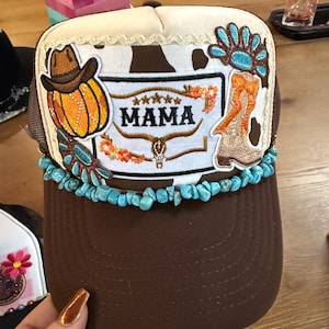 Peut inclure: Casquette de camionneur marron et beige avec une visière marron. Le chapeau présente un écusson avec le mot "MAMA" et une citrouille, une botte de cowboy et des accents turquoise. Le chapeau a une bordure en pierres turquoises.