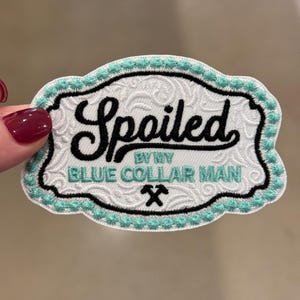 Puede incluir: Parche bordado con las palabras "Spoiled by my Blue Collar Man" en negro y verde azulado. El parche tiene un fondo blanco con un borde decorativo y un borde cosido en verde azulado. Un pequeño gráfico de martillos cruzados está en la parte inferior.