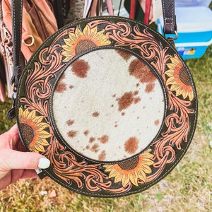 May include: A round, brown leather purse with a cowhide center and sunflower designs. The purse has a brown strap and is decorated with intricate floral patterns.