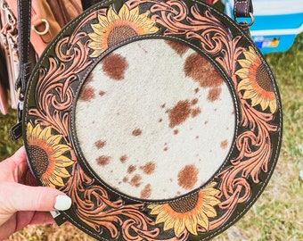 Op de afbeelding: Een ronde, bruine leren tas met een koeienhuid middenstuk en zonnebloemontwerpen. De tas heeft een bruine band en is versierd met ingewikkelde bloemmotieven.