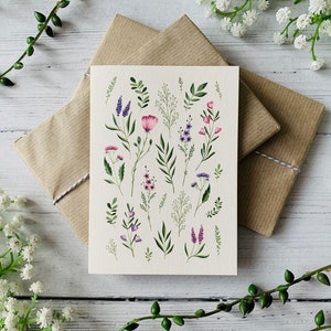 K&ouml;nnte beinhalten: Eine Karte mit einer Aquarell-Illustration verschiedener rosa und lila Blumen und gr&uuml;ner Bl&auml;tter auf cremefarbenem Hintergrund.