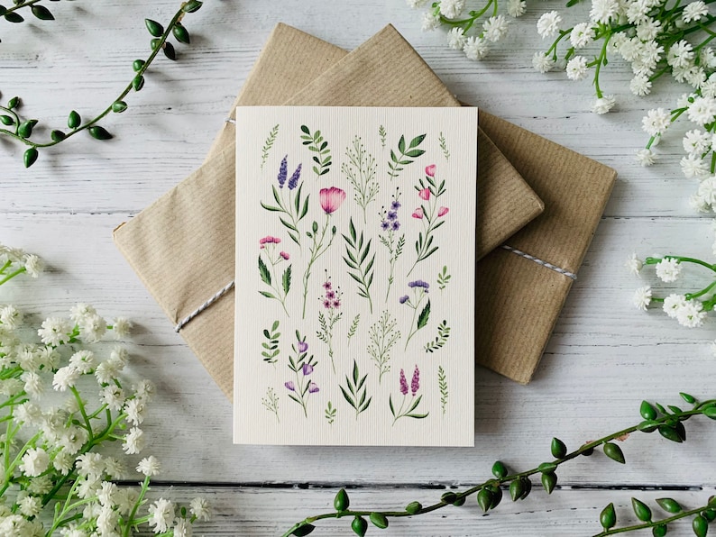 K&ouml;nnte beinhalten: Eine Karte mit einer Aquarell-Illustration verschiedener rosa und lila Blumen und gr&uuml;ner Bl&auml;tter auf cremefarbenem Hintergrund.