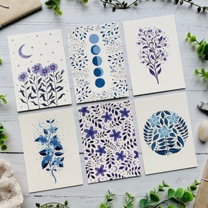 May include: Set of six illustrated cards featuring floral designs in shades of blue and purple. The cards feature different floral patterns, including a moon and stars, a circle of flowers, and a single branch of flowers.