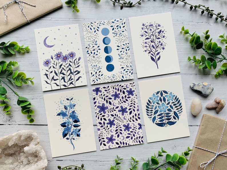 May include: Set of six illustrated cards featuring floral designs in shades of blue and purple. The cards feature different floral patterns, including a moon and stars, a circle of flowers, and a single branch of flowers.