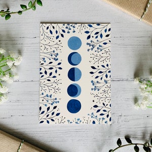 May include: A white card with a blue and black moon phase illustration surrounded by a border of blue and black floral designs.