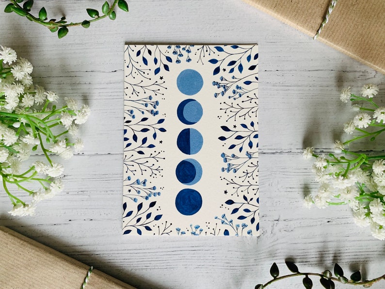 May include: A white card with a blue and black moon phase illustration surrounded by a border of blue and black floral designs.