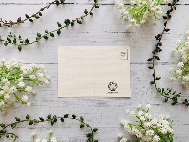 May include: A blank white postcard with a space for a stamp and an address. The postcard is surrounded by white flowers and green vines.