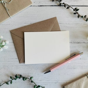 Puede incluir: Una tarjeta blanca en blanco con un sobre marr&oacute;n, un bol&iacute;grafo rosa y flores blancas sobre un fondo de madera blanco.