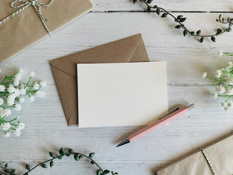 Puede incluir: Una tarjeta blanca en blanco con un sobre marr&oacute;n, un bol&iacute;grafo rosa y flores blancas sobre un fondo de madera blanco.