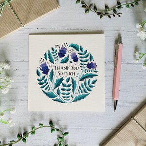 May include: A white card with a blue and green floral design and the text "Thank You So Much". The card is surrounded by white flowers, green plants, and a pink pen.