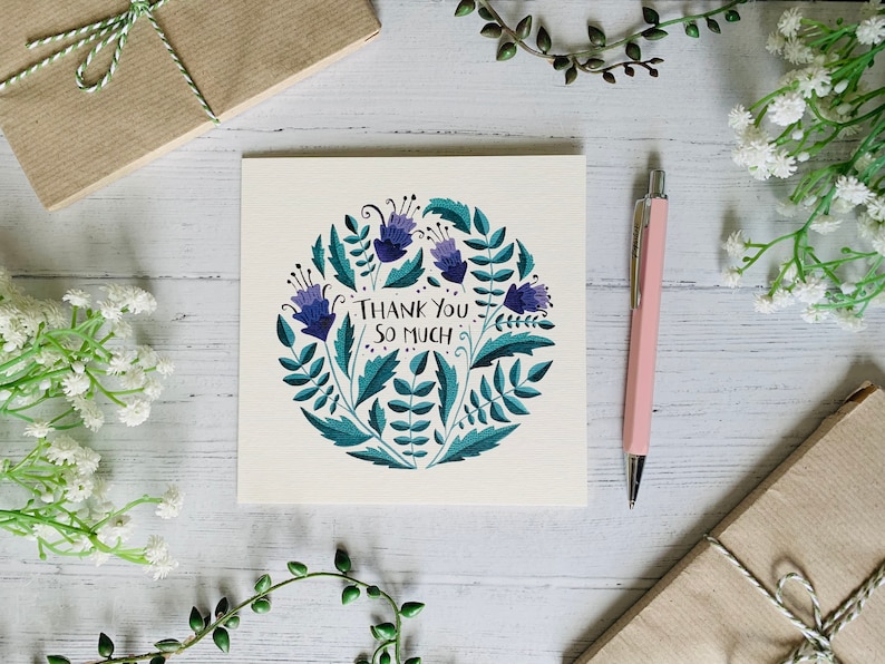 May include: A white card with a blue and green floral design and the text "Thank You So Much". The card is surrounded by white flowers, green plants, and a pink pen.