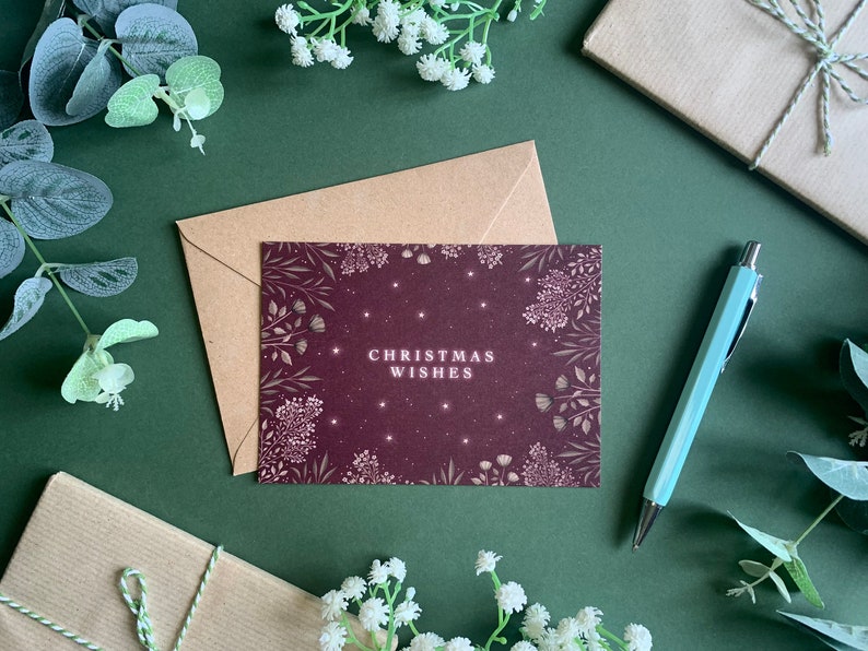 May include: A burgundy card with a white floral design and the text "Christmas Wishes" in white. The card is surrounded by greenery and white flowers.