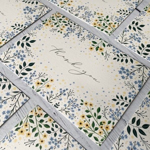 Puede incluir: Un mont&oacute;n de tarjetas de agradecimiento con un dise&ntilde;o floral con flores azules y amarillas sobre un fondo blanco. El texto "Gracias" est&aacute; escrito en cursiva negra.