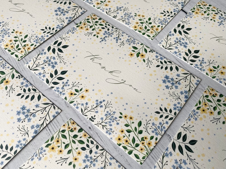 Puede incluir: Un mont&oacute;n de tarjetas de agradecimiento con un dise&ntilde;o floral con flores azules y amarillas sobre un fondo blanco. El texto "Gracias" est&aacute; escrito en cursiva negra.