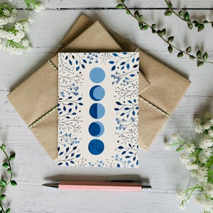 May include: A white card with a blue and white floral design and a row of blue crescent moons. The card is sitting on a brown envelope.