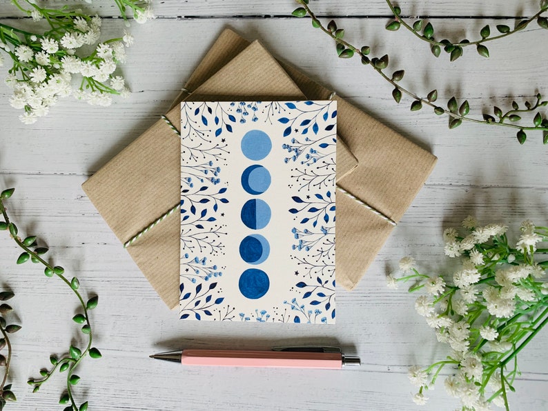 May include: A white card with a blue and white floral design and a row of blue crescent moons. The card is sitting on a brown envelope.