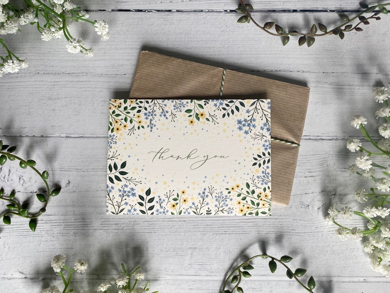 Puede incluir: Una tarjeta blanca con un borde floral en tonos azules y amarillos. La tarjeta dice "Gracias" en una fuente manuscrita.