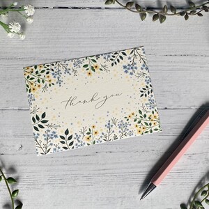 Puede incluir: Una tarjeta blanca con un borde floral y el texto "Gracias" en caligraf&iacute;a negra. La tarjeta est&aacute; sobre una superficie de madera blanca con un bol&iacute;grafo rosa.