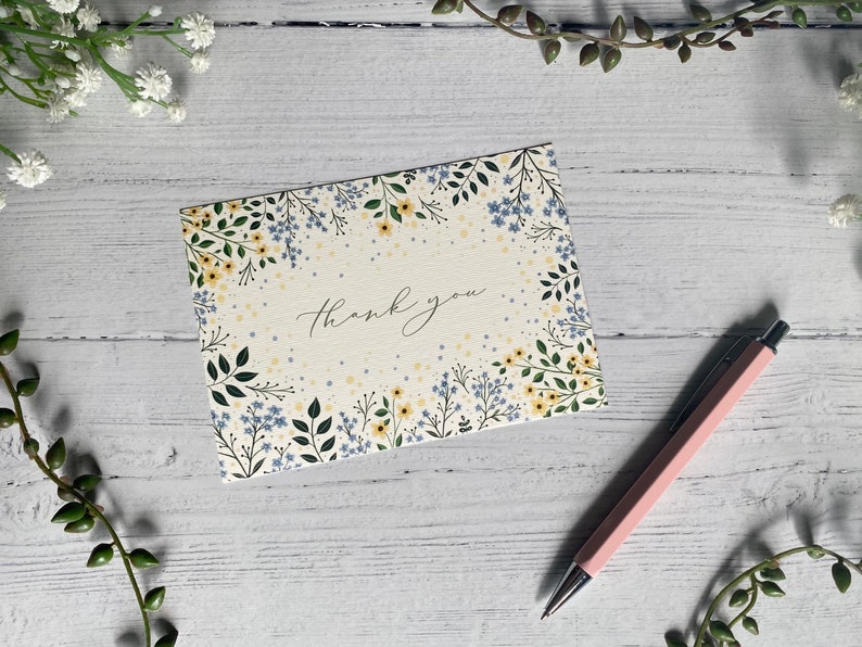 Puede incluir: Una tarjeta blanca con un borde floral y el texto "Gracias" en caligraf&iacute;a negra. La tarjeta est&aacute; sobre una superficie de madera blanca con un bol&iacute;grafo rosa.