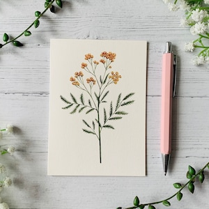 K&ouml;nnte beinhalten: Eine leere Karte mit einer zarten Illustration einer gelben Blume mit gr&uuml;nen Bl&auml;ttern. Die Karte liegt auf einer wei&szlig;en Holzoberfl&auml;che mit einem rosa Stift.