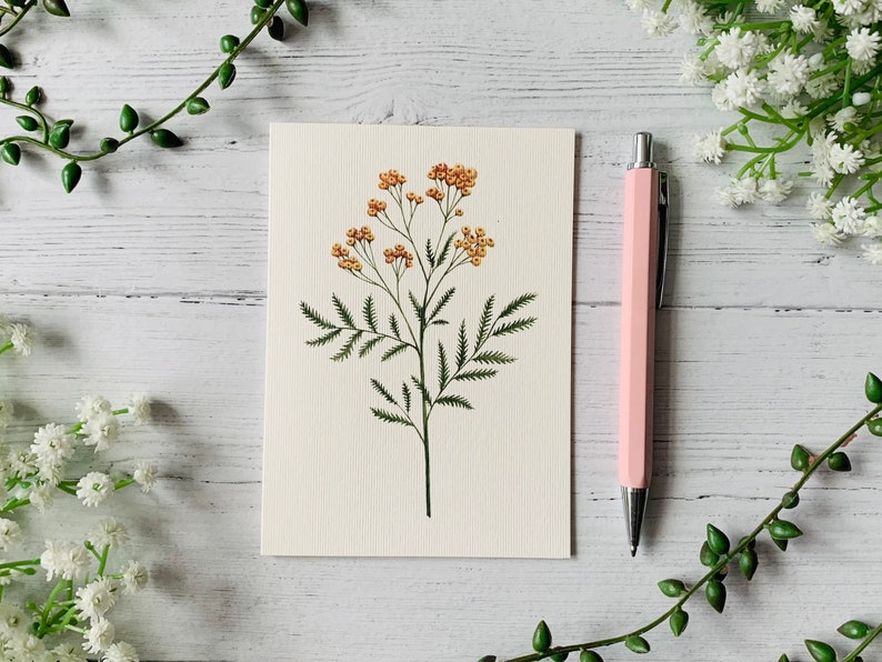 K&ouml;nnte beinhalten: Eine leere Karte mit einer zarten Illustration einer gelben Blume mit gr&uuml;nen Bl&auml;ttern. Die Karte liegt auf einer wei&szlig;en Holzoberfl&auml;che mit einem rosa Stift.