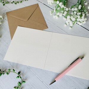 May include: A blank white card with a pink pen on a white wooden surface. The card is folded in half and has a brown envelope next to it. There are white flowers and green vines in the background.