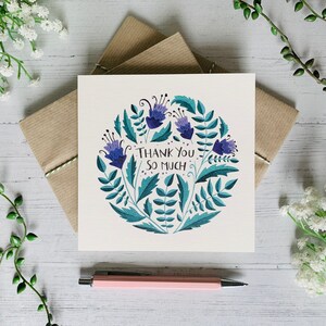 May include: A white card with a blue and green floral design and the text "Thank You So Much". The card is sitting on a white wooden surface with green plants and white flowers.