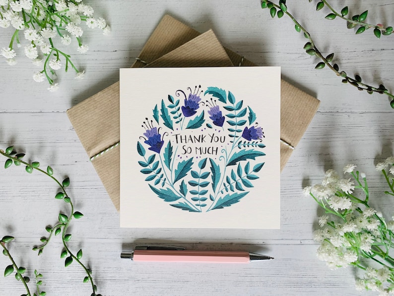 May include: A white card with a blue and green floral design and the text "Thank You So Much". The card is sitting on a white wooden surface with green plants and white flowers.