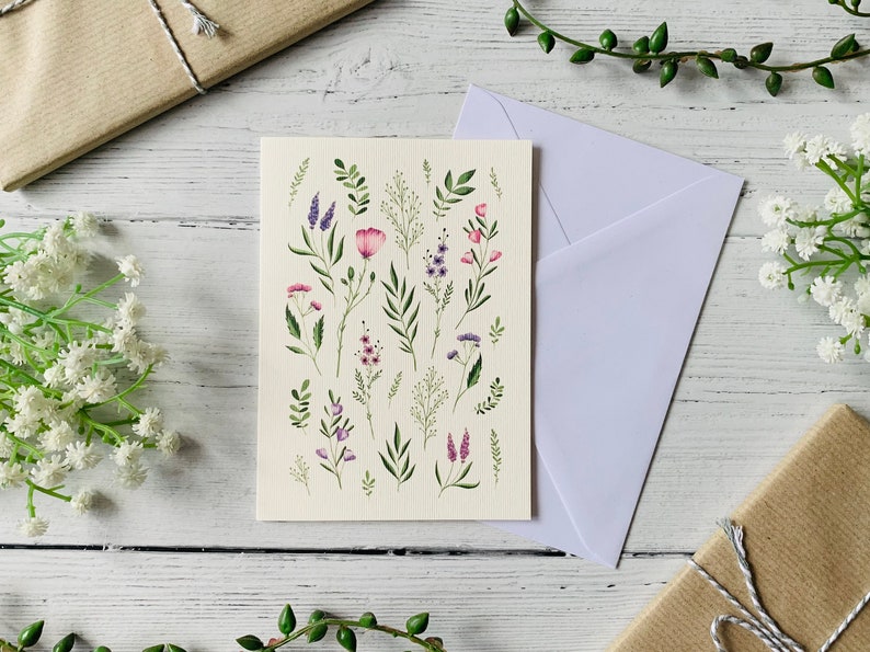 K&ouml;nnte beinhalten: Eine wei&szlig;e Karte mit einer Aquarell-Illustration von rosa und lila Blumen und gr&uuml;nen Bl&auml;ttern. Die Karte ist leer und bereit f&uuml;r eine Nachricht.