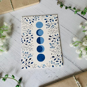 May include: A white card with a blue moon phase design. The card features a series of blue moons in different phases, surrounded by delicate black floral illustrations.