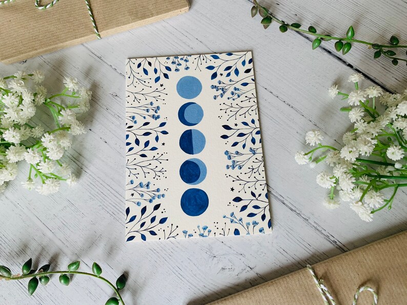 May include: A white card with a blue moon phase design. The card features a series of blue moons in different phases, surrounded by delicate black floral illustrations.