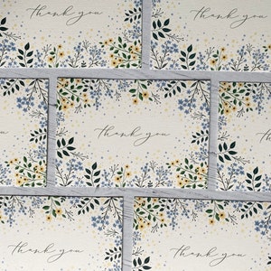 Puede incluir: Conjunto de seis tarjetas de agradecimiento con un dise&ntilde;o floral. Las tarjetas presentan un fondo blanco con flores y hojas azules y amarillas. El texto "Gracias" est&aacute; escrito en cursiva negra en cada tarjeta.