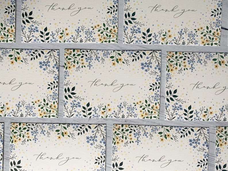 Puede incluir: Conjunto de seis tarjetas de agradecimiento con un dise&ntilde;o floral. Las tarjetas presentan un fondo blanco con flores y hojas azules y amarillas. El texto "Gracias" est&aacute; escrito en cursiva negra en cada tarjeta.