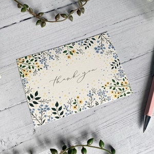 Puede incluir: Una tarjeta blanca con un borde floral y el texto "Gracias" en caligraf&iacute;a negra. Las flores son amarillas y azules.