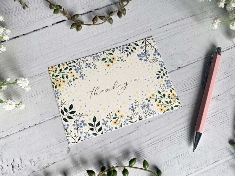 Puede incluir: Una tarjeta blanca con un borde floral y el texto "Gracias" en caligraf&iacute;a negra. Las flores son amarillas y azules.
