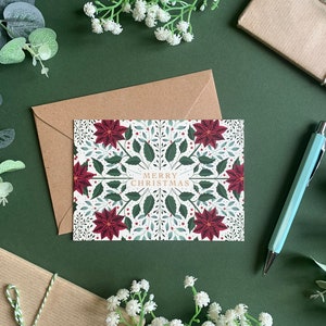May include: A white card with a green and red floral design and the text "Merry Christmas". The card is on a green background with brown paper envelopes and white flowers.