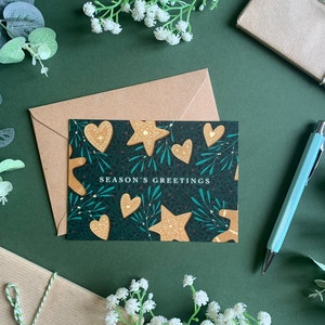 May include: A dark green card with a festive design featuring gingerbread cookies in the shape of stars and hearts. The card has the text "Season's Greetings" printed in white.