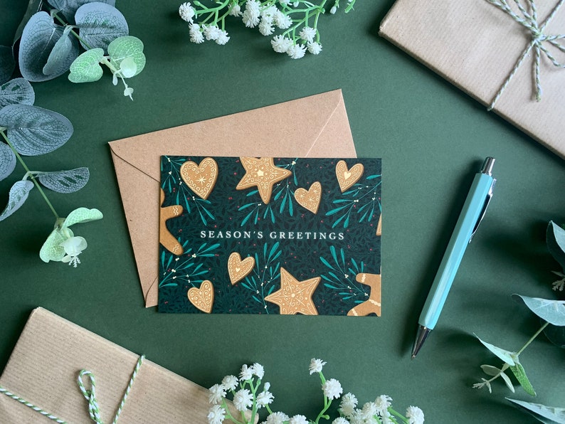 May include: A dark green card with a festive design featuring gingerbread cookies in the shape of stars and hearts. The card has the text "Season's Greetings" printed in white.