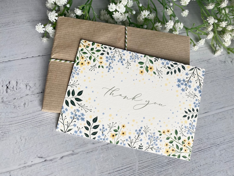 Puede incluir: Una tarjeta blanca con un borde floral de flores amarillas y azules. La tarjeta dice "Gracias" en escritura cursiva.