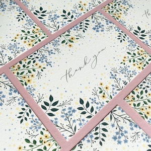 Puede incluir: Un conjunto de tarjetas de agradecimiento con un dise&ntilde;o floral con flores azules y amarillas sobre un fondo blanco. Las tarjetas tienen un borde rosa y el texto "gracias" est&aacute; escrito en cursiva.