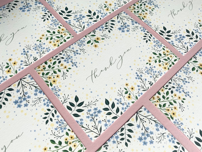Puede incluir: Un conjunto de tarjetas de agradecimiento con un dise&ntilde;o floral con flores azules y amarillas sobre un fondo blanco. Las tarjetas tienen un borde rosa y el texto "gracias" est&aacute; escrito en cursiva.