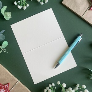May include: A blank white card with a light blue pen on top of it. The card is folded in half and is sitting on a green surface. There are sprigs of greenery and small white flowers around the card.