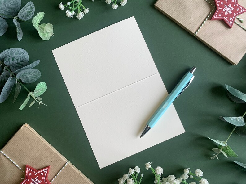 May include: A blank white card with a light blue pen on top of it. The card is folded in half and is sitting on a green surface. There are sprigs of greenery and small white flowers around the card.