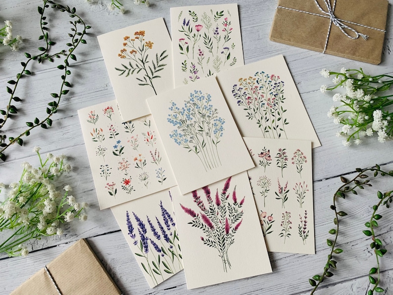 K&ouml;nnte beinhalten: Satz von acht Karten mit verschiedenen Blumenillustrationen. Die Karten sind wei&szlig; mit schwarzer Tinte und zeigen verschiedene Blumen, darunter blaue Vergissmeinnicht, rosa und violette Wildblumen, gelbe Wildblumen und Lavendel.