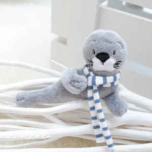 以下が含まれることがあります： 青と白のストライプのストールを巻いた灰色のぬいぐるみアザラシ。アザラシは白い枝の山の上に横たわっています。